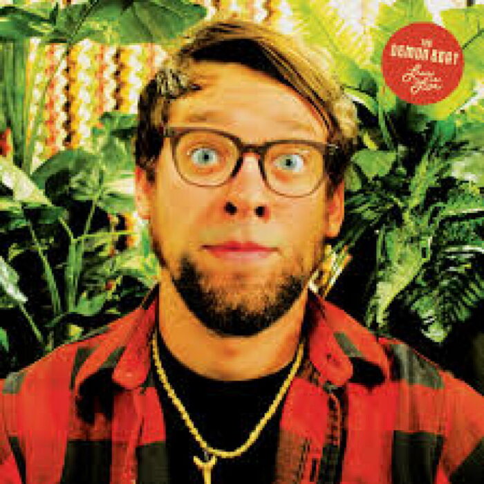 A photo of a man in his 20s or 30s. He wears glasses and looks toward the camera eyes wide. He's wearing a red plaid shirt. The image appears to be an album cover.