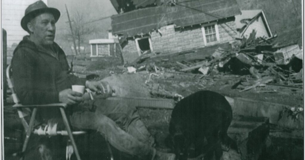 A black and white photo of a man sitting in a chair outside a house.