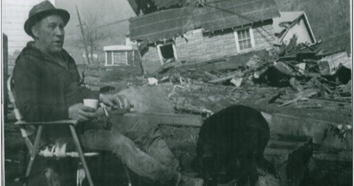 A black and white photo of a man sitting in a chair outside a house.