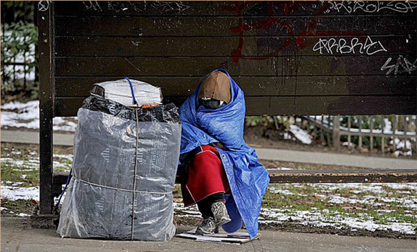 A photograph of someone who is homeless and sitting on the side of a street. They are wrapped up in a garbage bag.