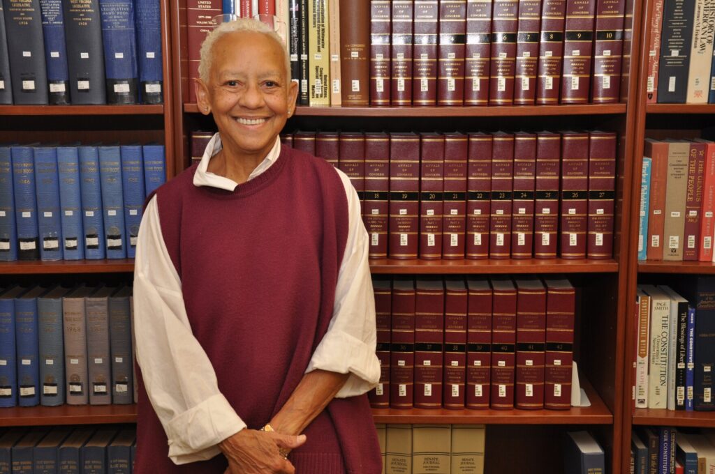 An older Black woman with short white hair stands against a bookshelf smiling. She wears a maroon sweater.