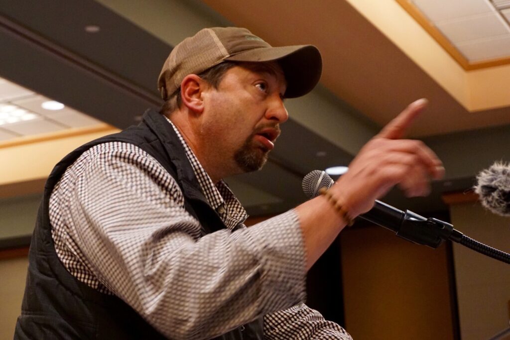 Organic farmer Shawn Peebles speaks into a microphone. He wears a ball cap and points his finger out toward the audience.