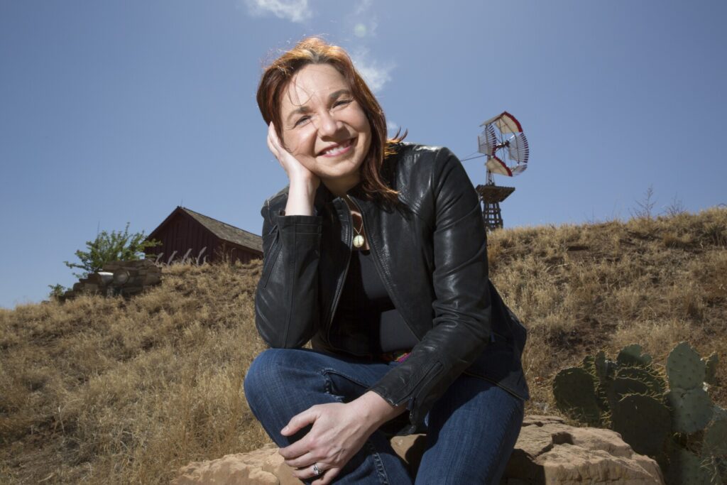 An adult woman smiles and poses for a photo. She sits on the side of a mountain. Above her is a blue sky. She wears a black jacket and blue jeans.