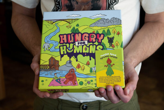 A man's hands hold a board game box that reads "Hungry for Humans."