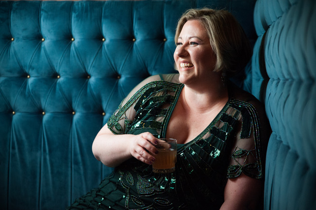 A woman dressed in a green dress, smiles while holding a glass, and leans against a blue cushioned wall.