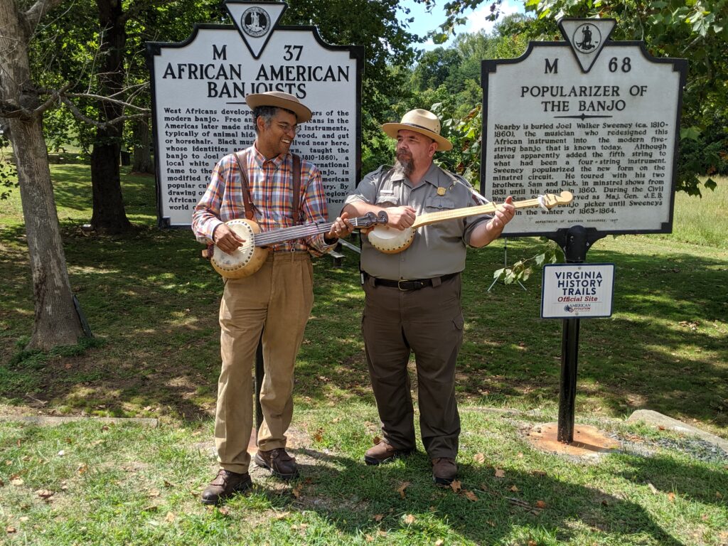 Two men stand next to each other playing banjos.