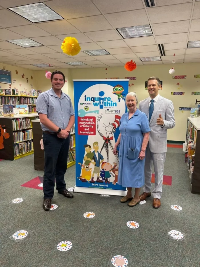 (L-R): Previous WVPB education specialist, Harrison Evans, Cabell County Public Library director, Judy Rule, and West Virginia state treasurer, Riley Moore, pose with the Inquire Within banner at the Cabell County Public Library 