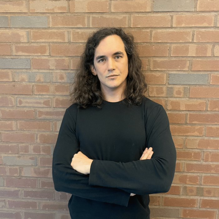 A man with long dark hair leans against a brick wall and looks at the camera.
