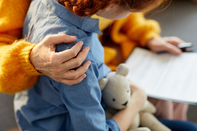 An adult embraces a child holding a teddy bear.