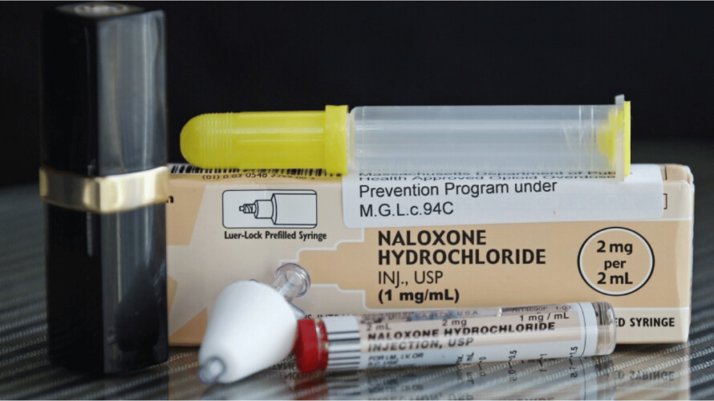 A syringe lays on top of a box with the words Naloxone written on it