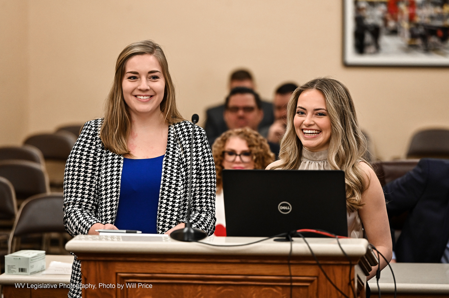 Two young women, blonde and smiling, stand side-by-side at a podium wearing formal attire in a West Virginia Senate committee room.