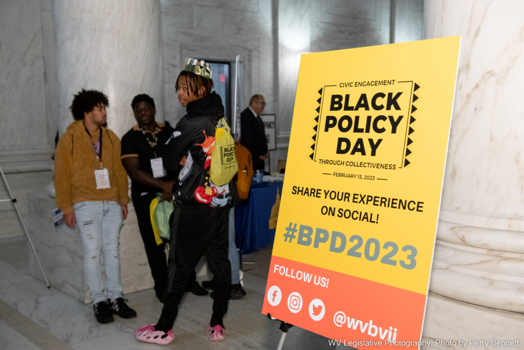 A group of young men, one wearing a gold crown, stand next to a yellow sign with a red band that reads, in part "Black Policy Day, Civic Engagement Through Collectiveness."
