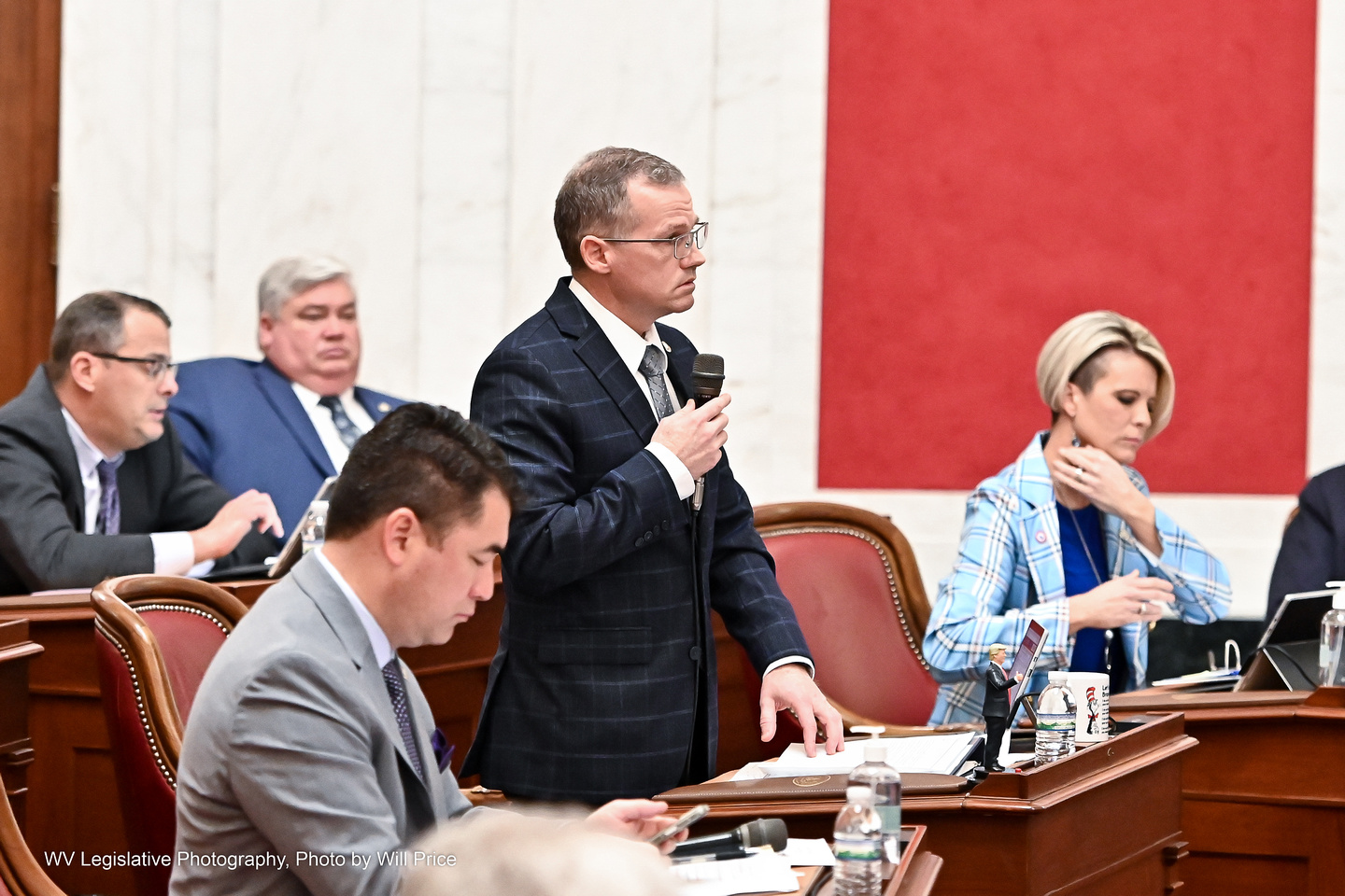 Sen. Eric Tarr stands on the Senate floor wearing a dark suit and holding a microphone up to speak while others around him sit