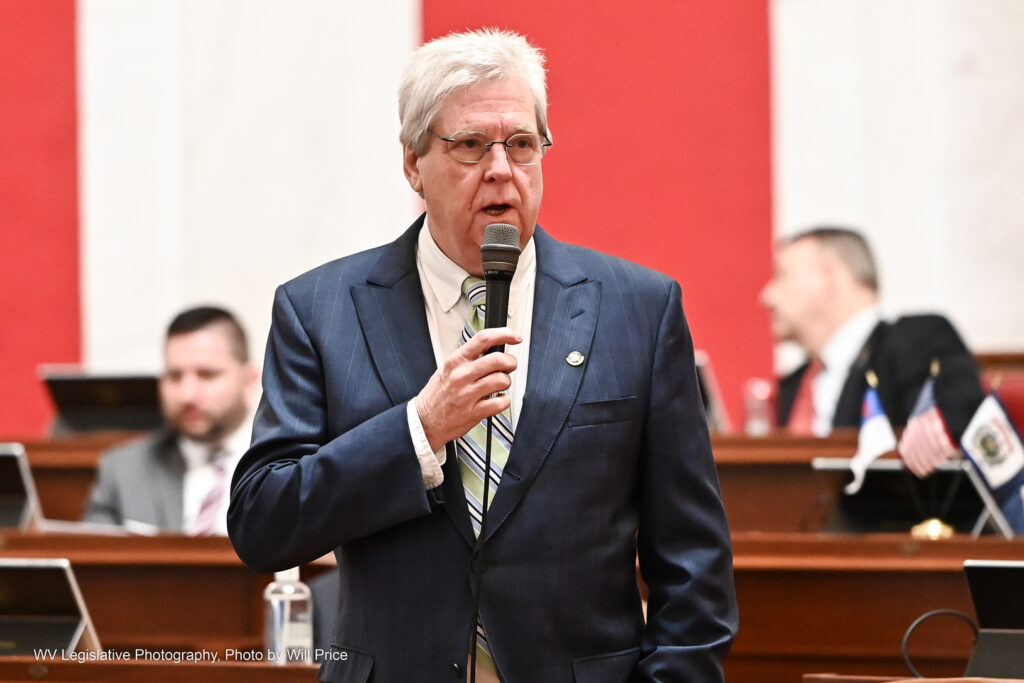 Sen. Mike Woelfel, wearing a mint green striped tie over a white dress shirt and a navy blue suit jacket and glasses, speaks on the Senate floor Feb. 22, 2023. Two other senators can be seen behind him at their desks.