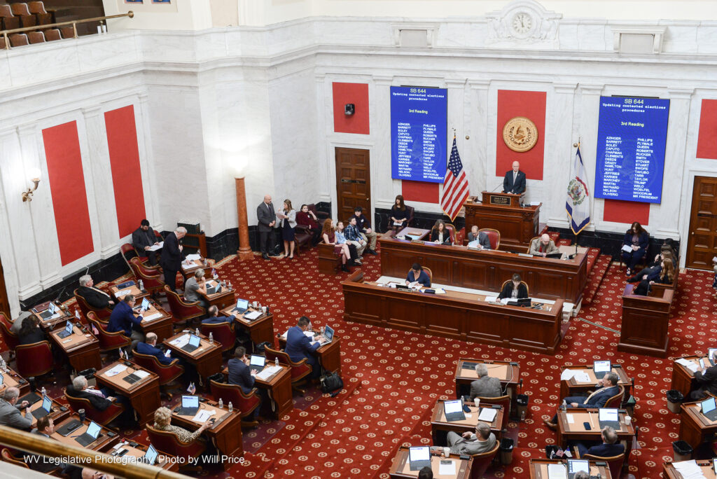 A view of the Senate chamber as seen from the President's Gallery shows dozens of people sitting at individual desks facing a dais. On blue screens, on either side of the dais, is displayed the chamber's voting roll for SB 644, titled "Updating contested elections procedures."