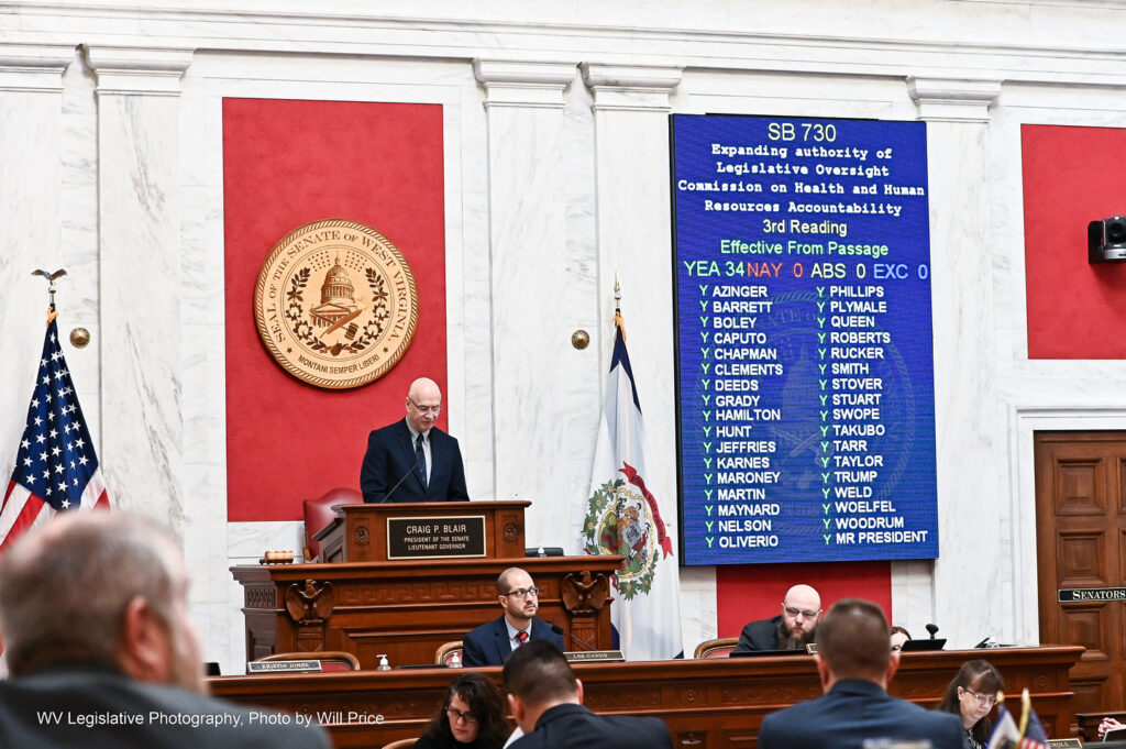 Senate President Craig Blair can be seen standing at the dais in the Senate Chamber. Next to him, the unanimous results passing Senate Bill 730, are displayed on a large, blue screen.