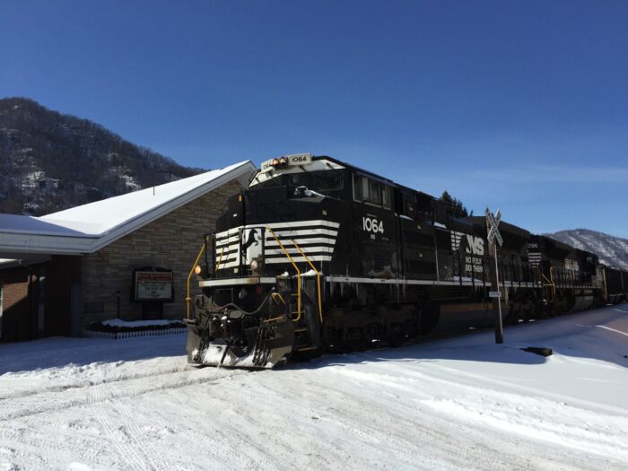 A Norfolk Southern coal train passes a small church in Boomer, West Virginia, with snow covering the ground and roof against a blue sky.