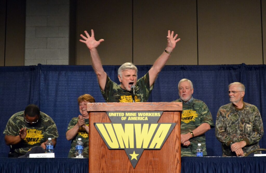 UMWA President Cecil Roberts, dressed in a camouflage shirt and surrounded by union members, waves a victory sign.