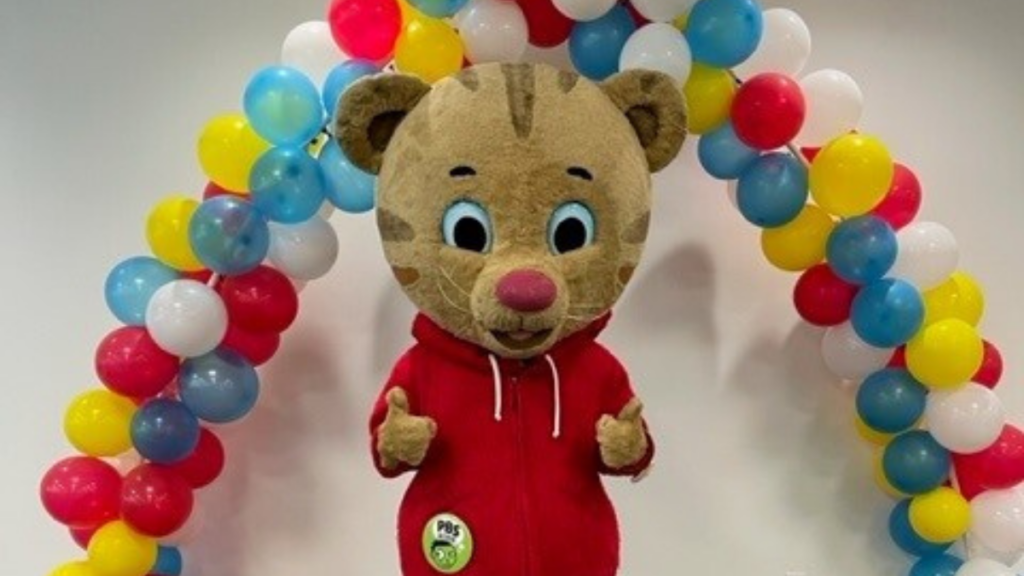 Bill Lynch dressed as Daniel Tiger poses for a photo under a balloon arch and gives two thumbs up.