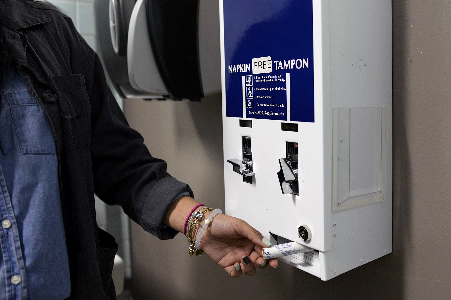 A woman reaches for a free tampon from a dispenser in a school restroom.