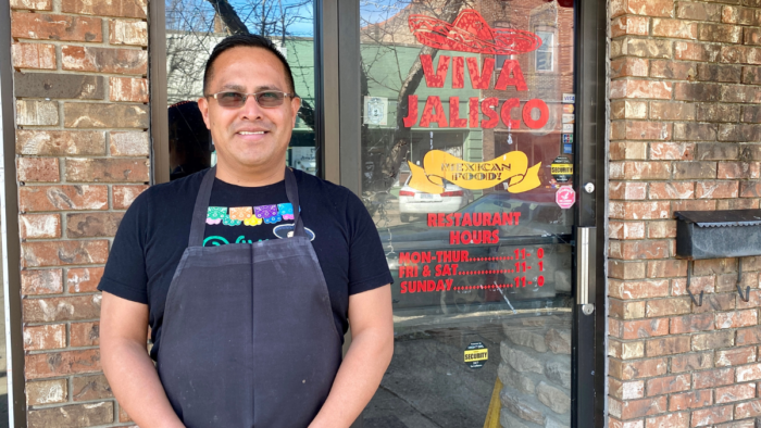 A man wearing glasses in a black shirt and dark gray apron stands smiling in front of a brick building with a glass door.
