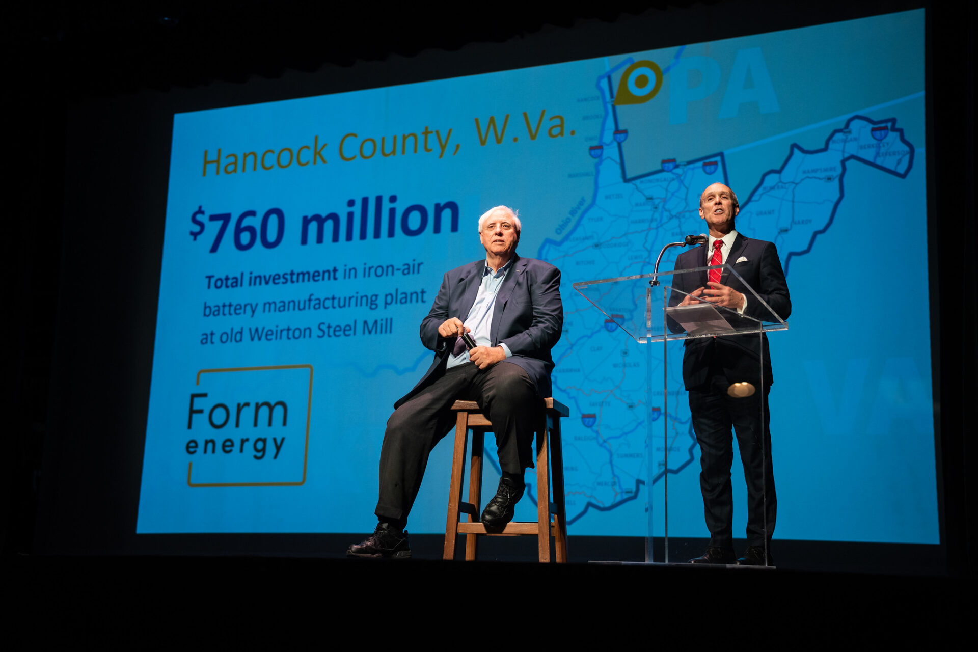 Two men in suits making an announcement in front of a large white sign with blue and yellow writing.