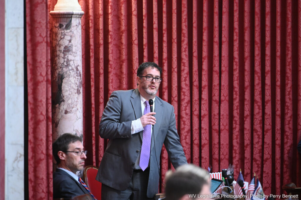 A man wearing a purple tie stands in the House of Delegates and speaks into a microphone.