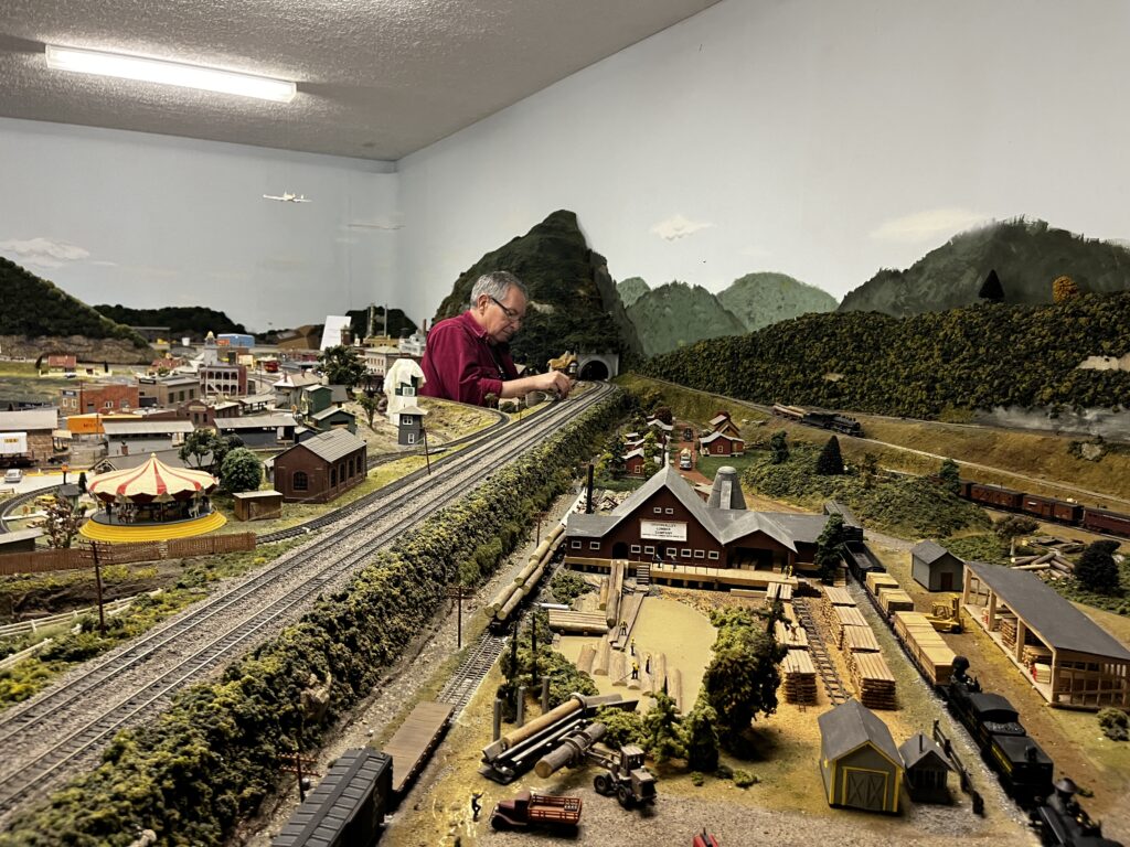A man sits at a table and works on a large model train and model town.