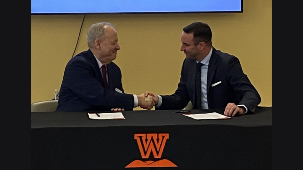 Two men are sitting and shaking hands behind a table emblazoned with West Virginia Wesleyan College's logo.