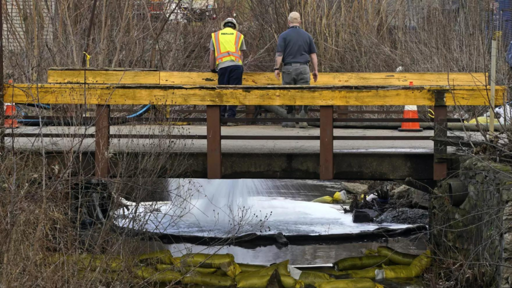 Two people peer over a bridge crossing a river that is under observation for chemical contamination.
