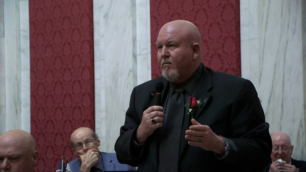 Delegate Scot Heckert, wearing a black suit with a rose lapel, speaks into a microphone on the House floor.