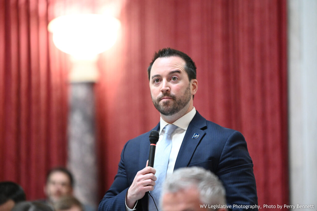 A man wearing a suit and tie stands holding a microphone in the West Virginia House of Delegates chamber. In the background, there is red cloth against a marble wall and a large lamp is turned on and glowing.