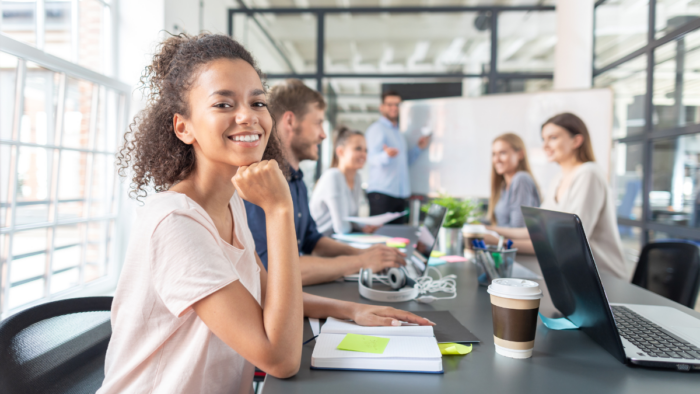 A young female entrepreneur smiles at the camera. She is sitting at a table full of other young people in what looks like a business meeting.