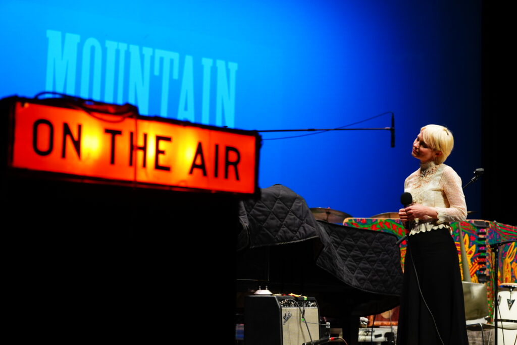 A short-haired, blonde woman stands on a stage dressed in a white shirt and black skirt. There is an "On The Air" light lit up and a screen up stage that reads "Mountain Stage."