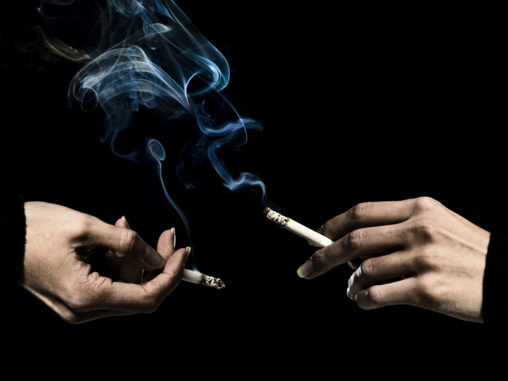 Two hands are seen smoking cigarettes with smoke standing stark against a black background.