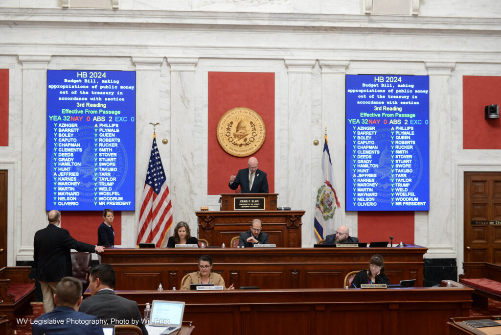 Senate President Craig Blair stands on the Senate dais between the completed vote tally for House Bill 2024, the state budget bill. The blue screens show 32 yeas, 0 nays and 2 absent.