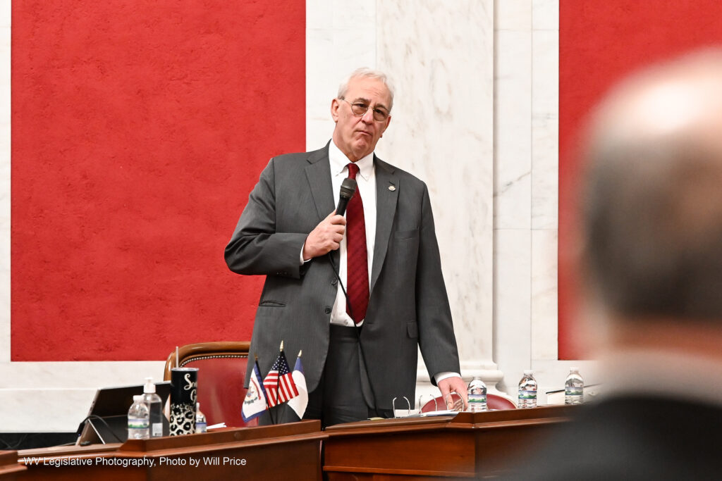 Sen. Charles Trump, R-Morgan, answers questions about a bill on the Senate floor Friday night, March 10, 2023. The background behind him is red to his left, and white marble to his right with a sliver of more red. Trump is wearing a grey suit over a red tie and white shirt. On the far right of the frame, the outline of another speaker's head can be seen in the extreme foreground.