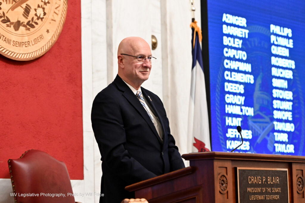 Senate President Craig Blair, R-Berkeley, wearing glasses, a black suit over a white shirt with a tan and light-blue striped tie, looks out over the Senate Chamber on March 11, 2023, the final night of the 2023 legislative session, as staff members are recognized for their work. To his left can be seen a portion of the Senate seal on a red background, and to his right the names of Senators are displayed on a blue screen.