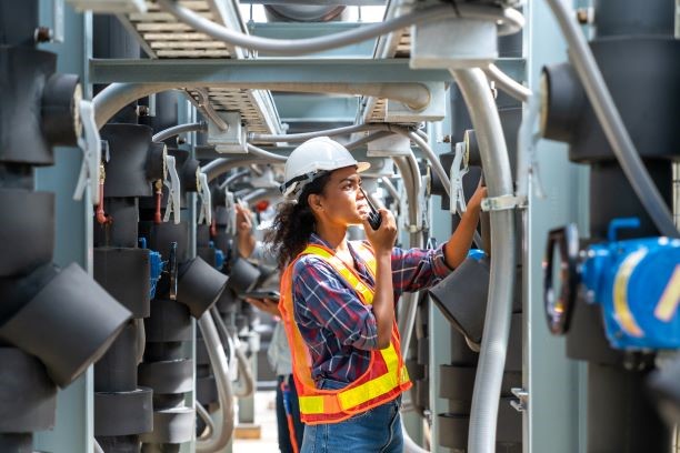 A woman wearing a high visibility vest and white hardhat speaks into a radio device while inspecting piping.