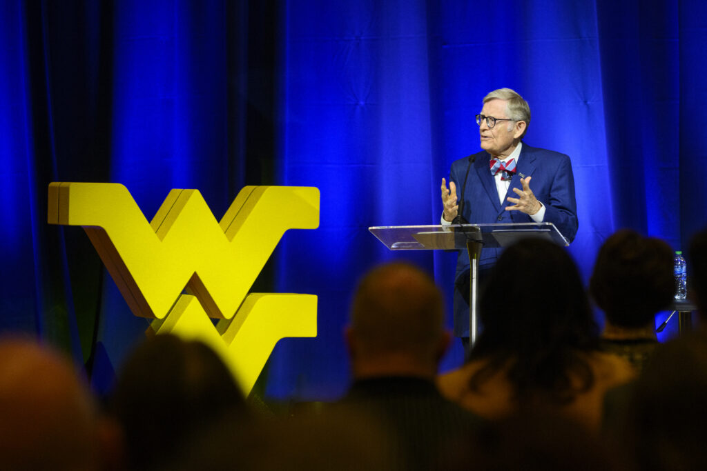 WVU President Gordon Gee stands in front of a blue curtain next to a large 'WV' logo statue. Gee wears a light red and blue bowtie over a white dress shirt and blue suit. The heads of the crowd he addresses can be seen in the foreground.