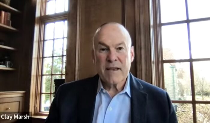 Coronavirus Czar Dr. Clay Marsh sits in front of large windows set in a wood-paneled wall. He wears a light blue shirt under a dark blue blazer.