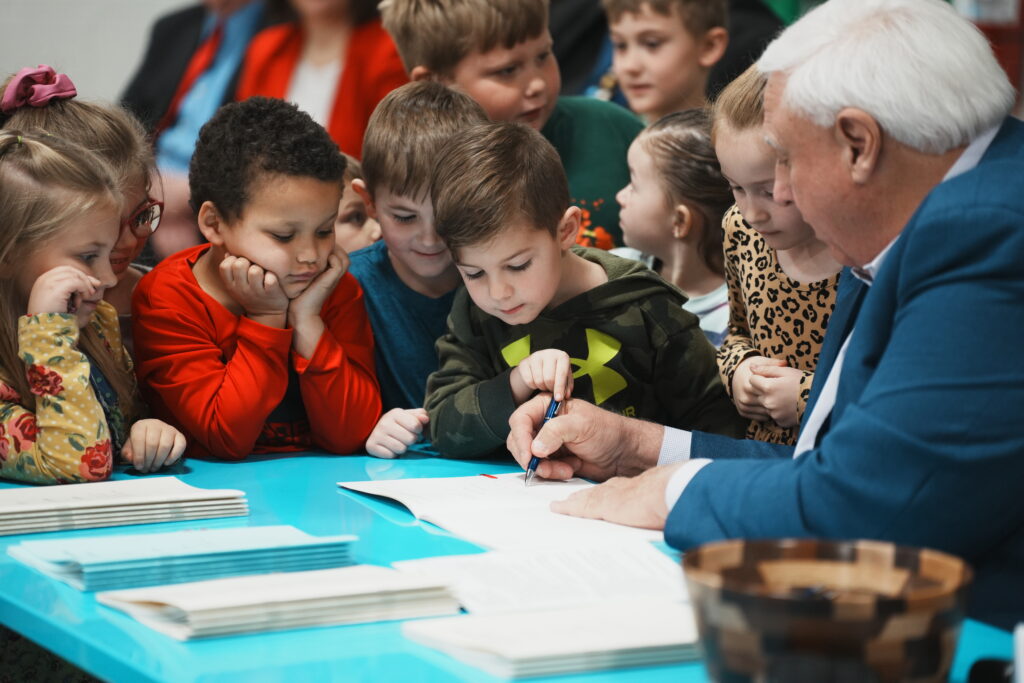 Big man with white hair at desk surrounded by grade schoolers.