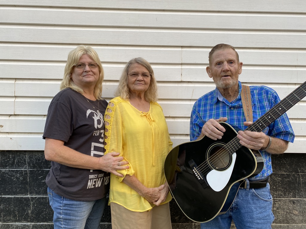 Three people stand side by side. One woman wears a black-grey shirt, one woman wears a yellow shirt, and a man in a blue shirt holds a black acoustic guitar.