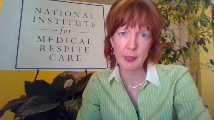 A middle age woman with red hair looks at the camera. She wears a light green work jacket. Behind her is a sign that reads, "National Institute for Medical Respite Care."