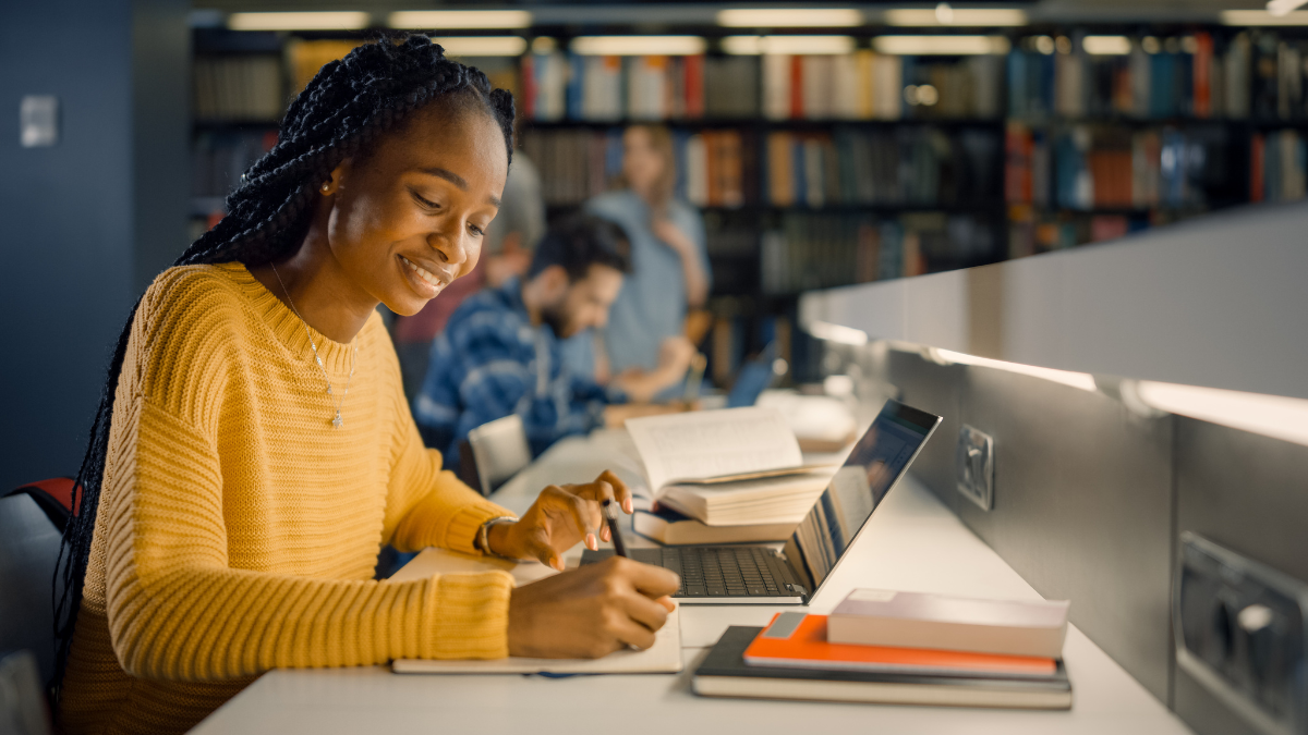 Black female student studying and smiling wearing yellow long sleeve shirt. She sits at a desk in a library.