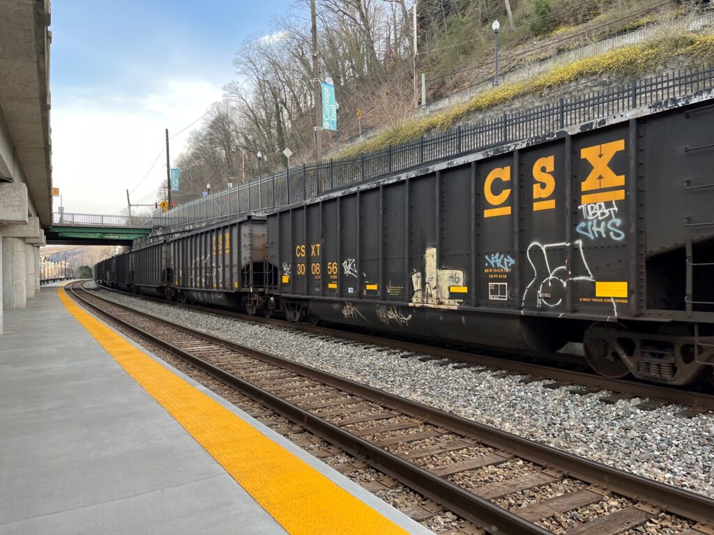 A CSX coal train ,with black cars and yellow letting, passes the concrete station platform at Charleston on an early spring day.