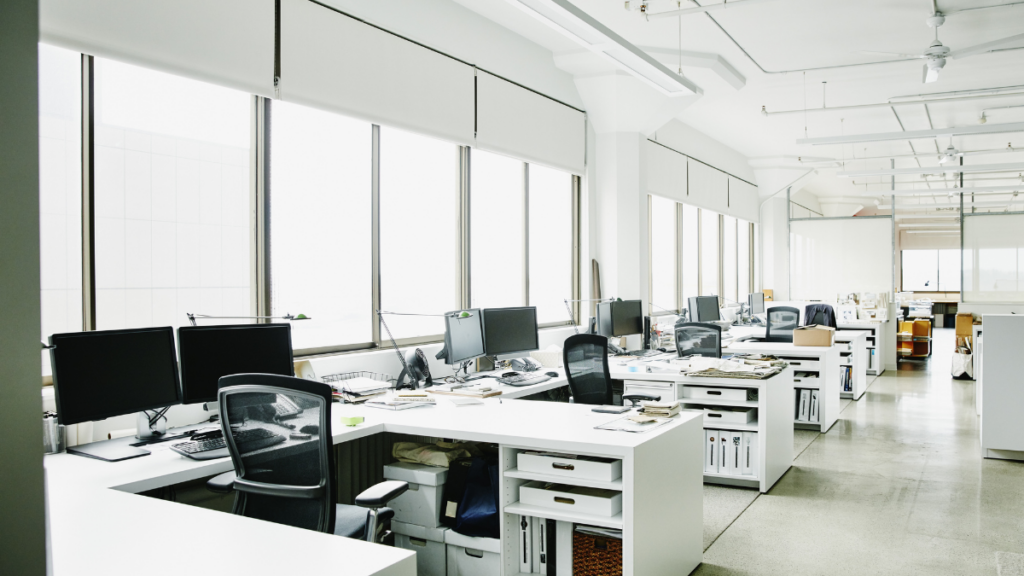 An open concept office shows desks up against high windows. Diffused, white light comes into the room through the windows, which is primarily white and beige tones.