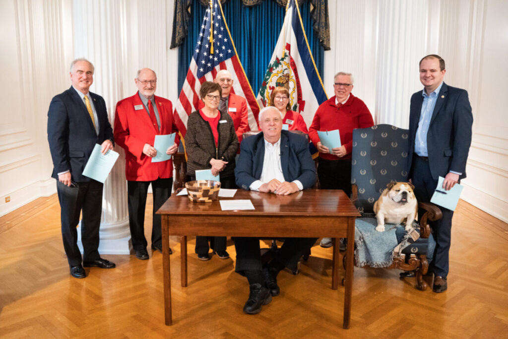 Seven people wearing formal attire stand behind the governor of West Virginia, Jim Justice. The governor's dog, a bulldog named Babydog, sits next to the governor in a chair.
