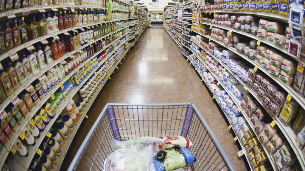 A grocery store aisle is pictured with a wide lens, showing both sides and the photographer's grocery cart.
