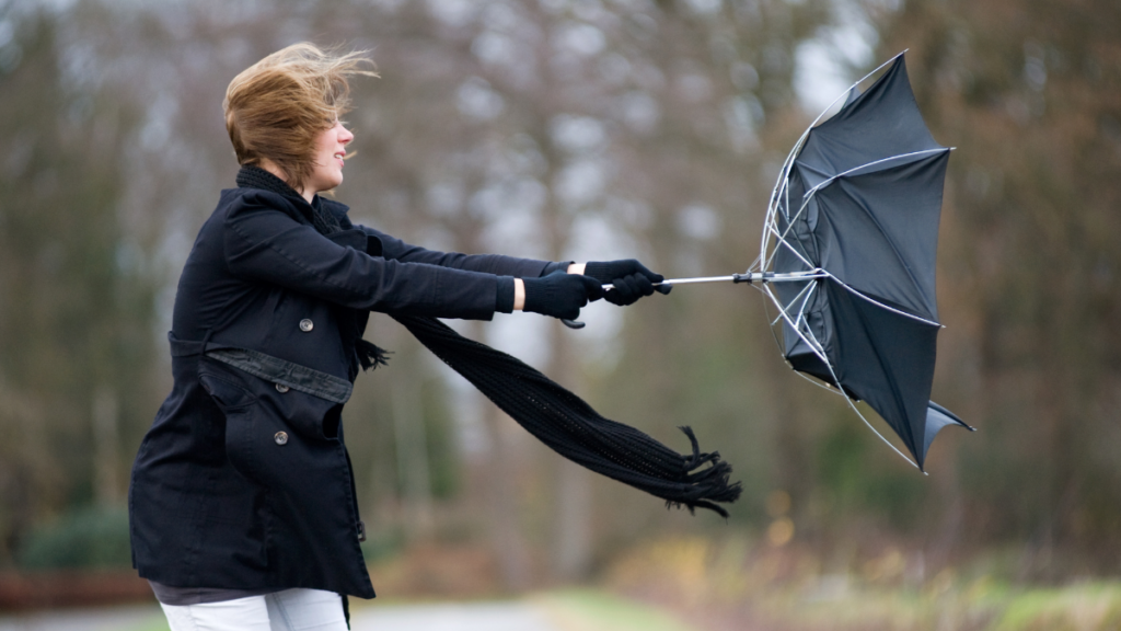 A woman wearing a black jacket and black gloves struggles with a black umbrella that has been flipped inside-out by strong winds. The woman's red hair is also being blown about.